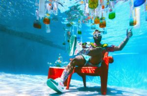 A young man in white-framed sunglasses sits on a red lawn chair next to a red cooler at the bottom of a pool with many bottles filled with different coloured drinks/liquids floating at the surface of the pool above him